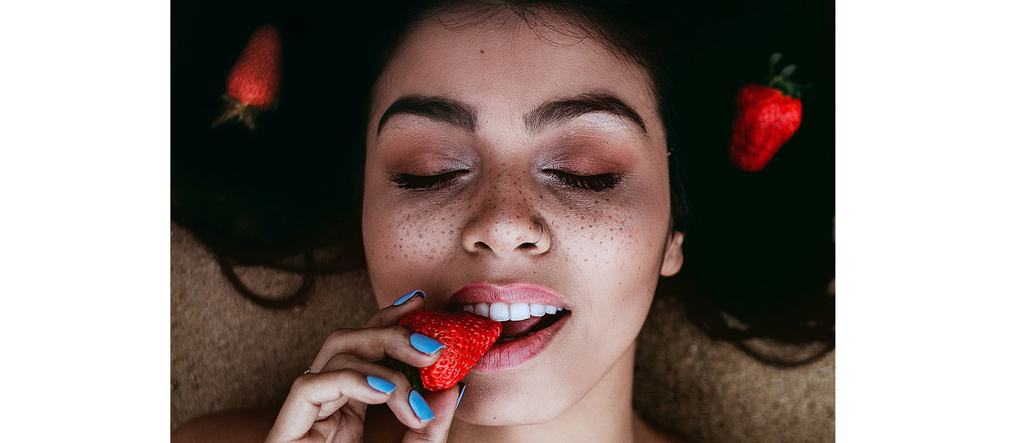 A close-up of a relaxed woman enjoying a strawberry, with a focus on her freckled face and vibrant red lips